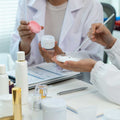 Two people in lab coats examining a cosmetic cream sample at a table with skincare containers, paperwork, and a laptop.