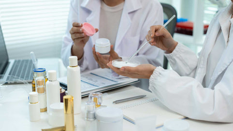Two people in lab coats examining a cosmetic cream sample at a table with skincare containers, paperwork, and a laptop.