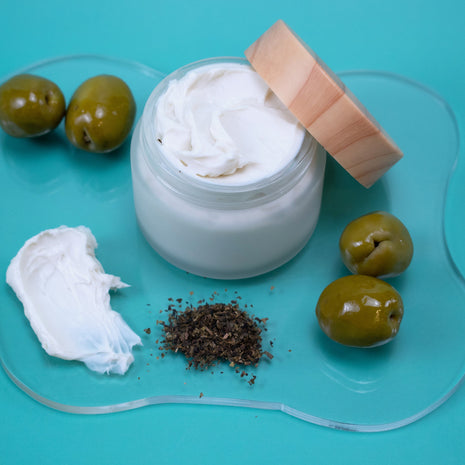 Jar of white cream with a wooden lid, surrounded by green olives on a blue background