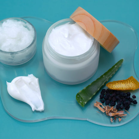 Jar of white cream with a wooden applicator, surrounded by natural ingredients on a blue background
