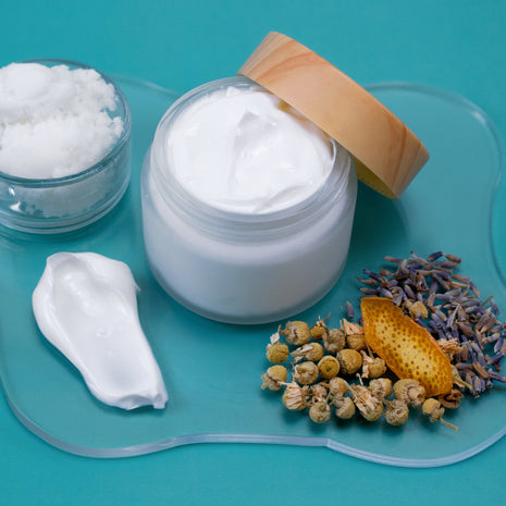 Jar of white cream with a wooden lid, surrounded by dried flowers and herbs on a blue background