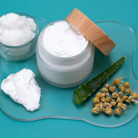 Jar of white cream with a wooden lid, surrounded by aloe vera leaf, dried flowers, and a small bowl on a blue background.