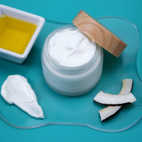 Jar of white cream with a wooden applicator, coconut pieces, and a small container of oil on a blue background.