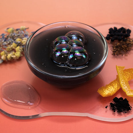 Glass bowl with dark liquid and soap bubbles on a pink surface with dried herbs and lemon peel.
