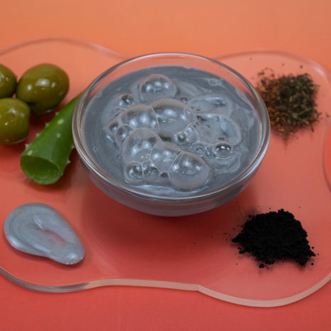 Gray gel in a clear container with green leaves and black powder on an orange background