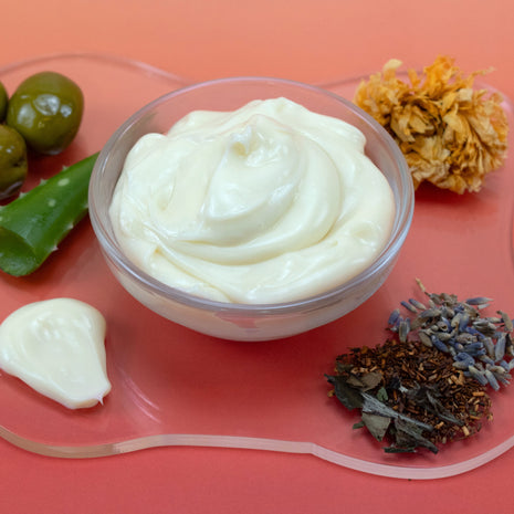 White cream in a clear container on a pink surface with green leaves, yellow flowers, and dried herbs.