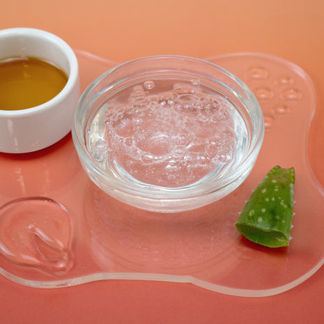 Clear container with liquid, white bowl with yellow substance, and green leaf on a pink surface