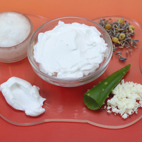 White cream in a glass bowl with aloe vera leaf, dried flowers, and salt on an orange background