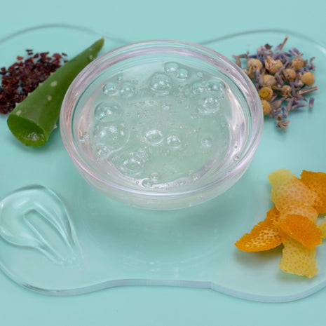 Clear gel in a container with aloe vera leaf, lavender flowers, and orange peel on a light blue background