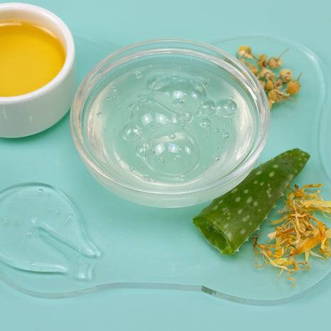 Clear gel in a small container with aloe vera leaf, calendula flowers, and a white bowl of yellow liquid on a light blue background.