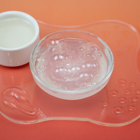 Clear silicone mold on a pink silicone mat with a small white bowl of liquid next to it on an orange background