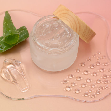 Jar of clear cream with aloe vera leaf and wooden applicator on a pink background