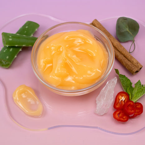 Muscle Gel in a glass bowl surrounded by aloe vera leaves, cinnamon sticks, and chili peppers on a pink background