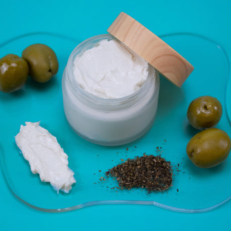 Olive and Green Tea Lotion with a wooden lid on a blue background with olives and other ingredients.