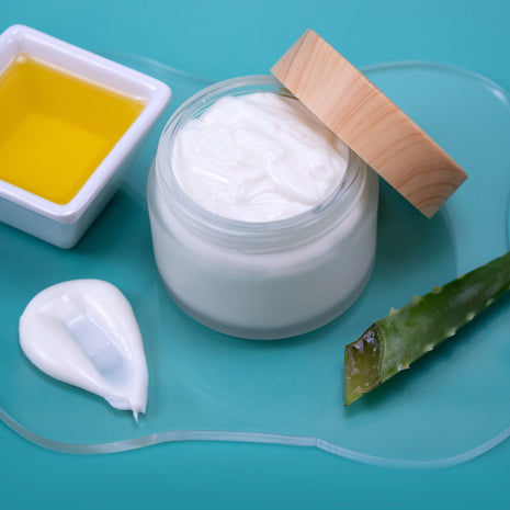 Aloe & Jojoba Lotion with a wooden lid, a small container of jojoba oil, and a piece of green aloe vera leaf on a teal background.
