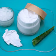 Basic Lotion with a wooden lid on a blue background, accompanied by aloe vera leaf and a spoonful of cream.