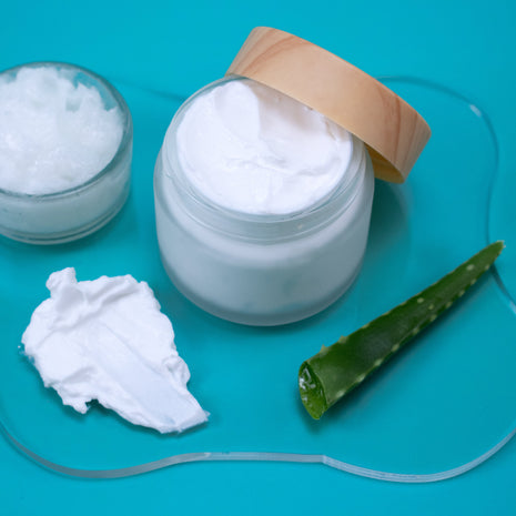 Basic Lotion with a wooden lid on a blue background, accompanied by aloe vera leaf and a spoonful of cream.