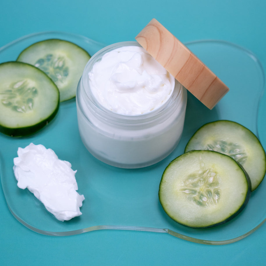 Jar of Cucumber Lotion with wooden lid, surrounded by sliced cucumbers on a blue background