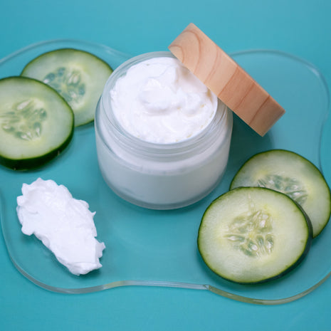 Jar of Cucumber Lotion with wooden lid, surrounded by sliced cucumbers on a blue background