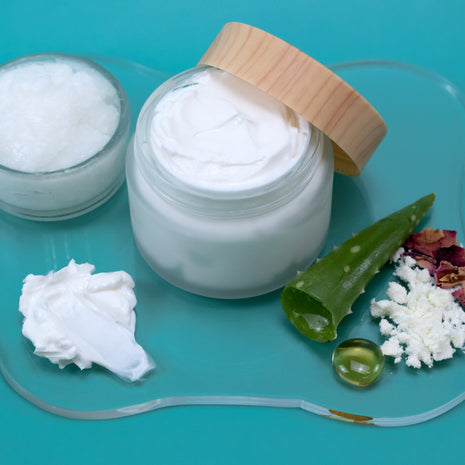 Jar of Goat Milk Lotion with a wooden lid on a teal background, surrounded by aloe vera leaves and other natural elements.