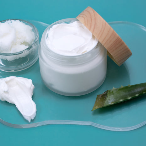 Jar of Shea Butter Lotion with a wooden lid on a teal background with aloe.