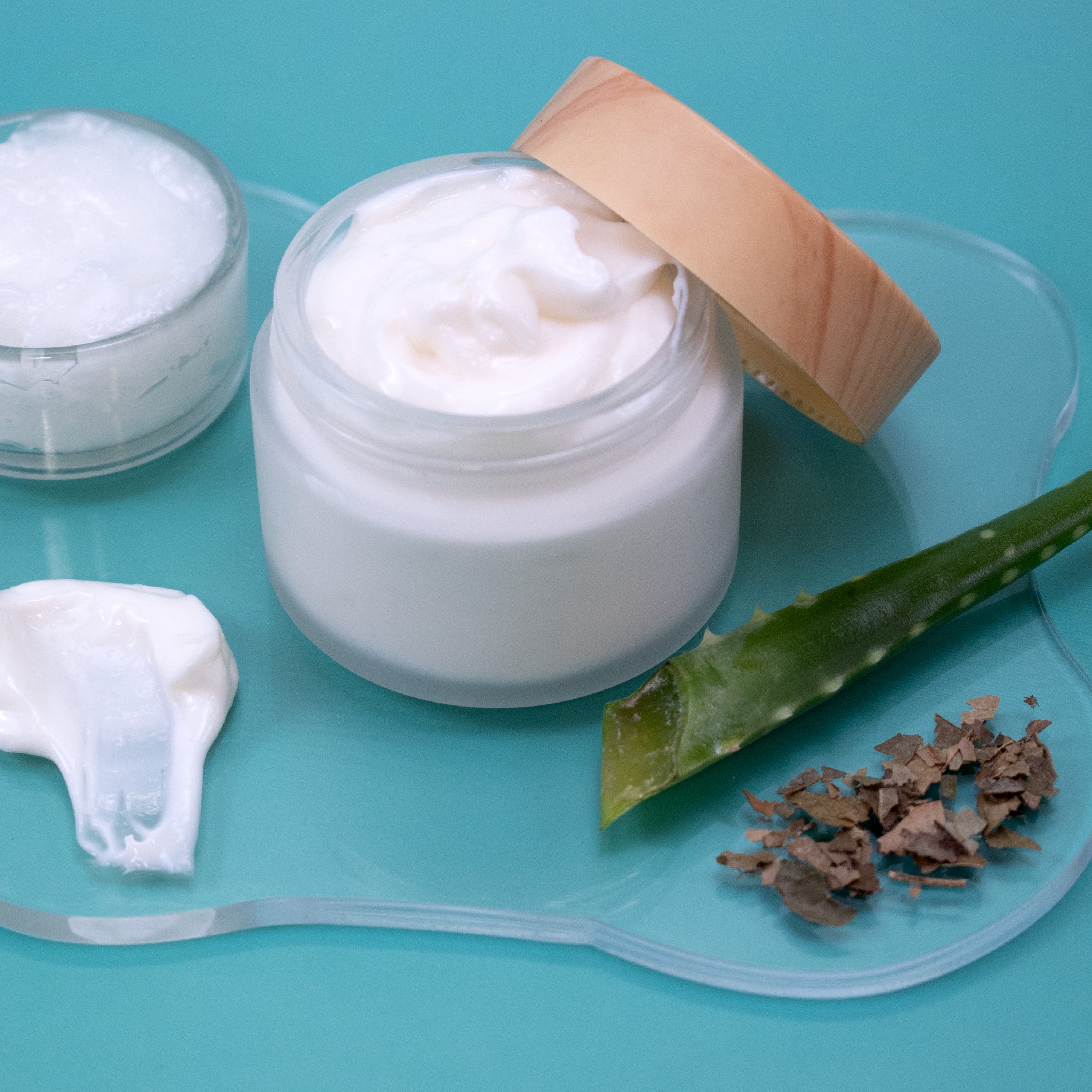 Jar of Soothing Lotion with a wooden lid on a blue background, surrounded by aloe vera leaves and pieces.