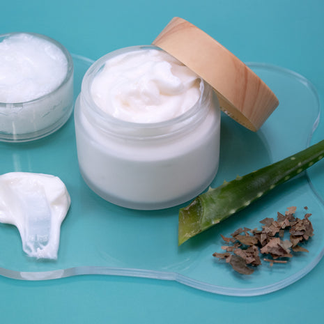 Jar of Soothing Lotion with a wooden lid on a blue background, surrounded by aloe vera leaves and pieces.