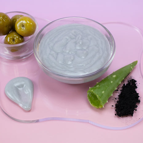 Glass bowl with Charcoal Cream Mask, aloe vera leaf, and charcoal powder on a pink background