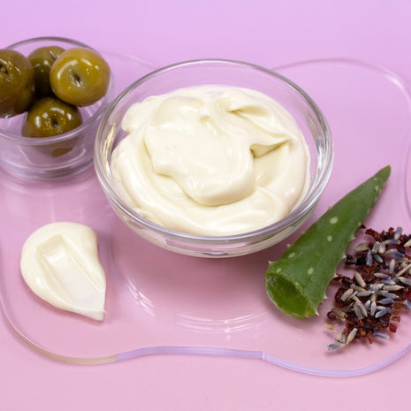 Bowl of Equalizer Cream Facial Mask with olives, aloe vera leaf, and lavender on a pink background