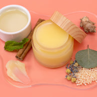 Jar of Muscle Rub with wooden lid on a pink background with natural ingredients