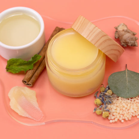 Jar of Muscle Rub with wooden lid on a pink background with natural ingredients