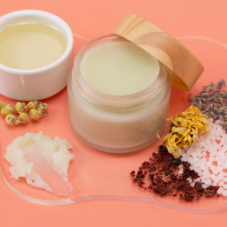 Body & Bum Balm jar with lid, small bowl, and various dried herbs on a peach-colored background
