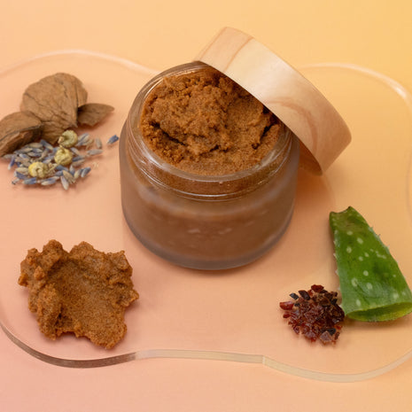 Glass jar with Walnut Body Polish, surrounded by dried herbs and aloe vera on a peach background