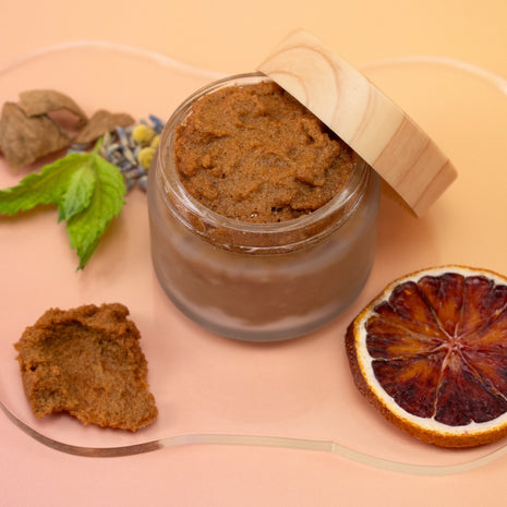 Jar of Blood Orange AHA Mint Walnut Body Polish with a wooden lid, surrounded by dried leaves and a blood orange slice on a peach background