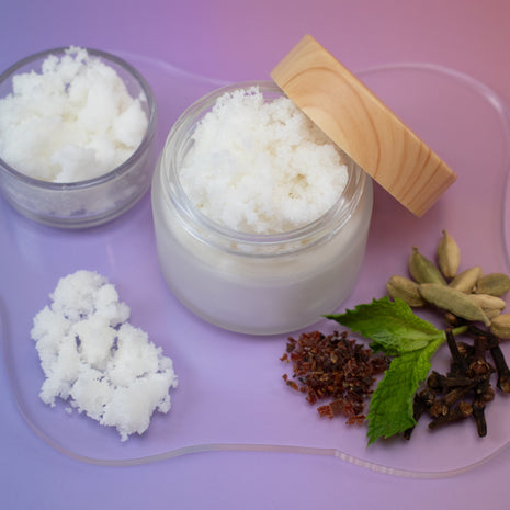 Jar of Autumn Aura Sugar Scrub with wooden lid on a pink tray with various herbs and spices.