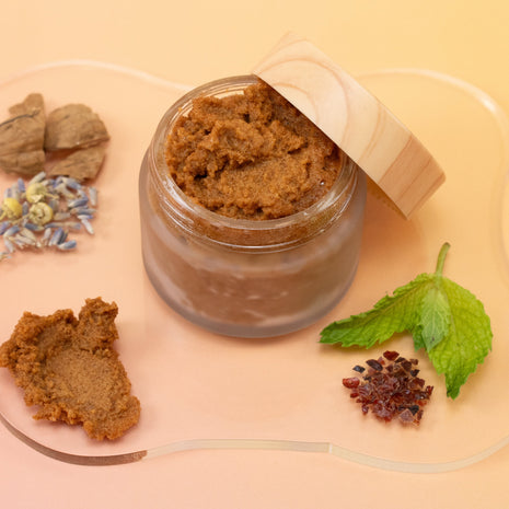 Glass jar with AHA Mint Walnut Body Polish, surrounded by dried herbs and leaves on a light pink background