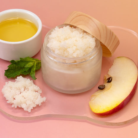 Jar of Love Your Body Scrub with a wooden lid, apple slices, and a small bowl of oil on a pink background
