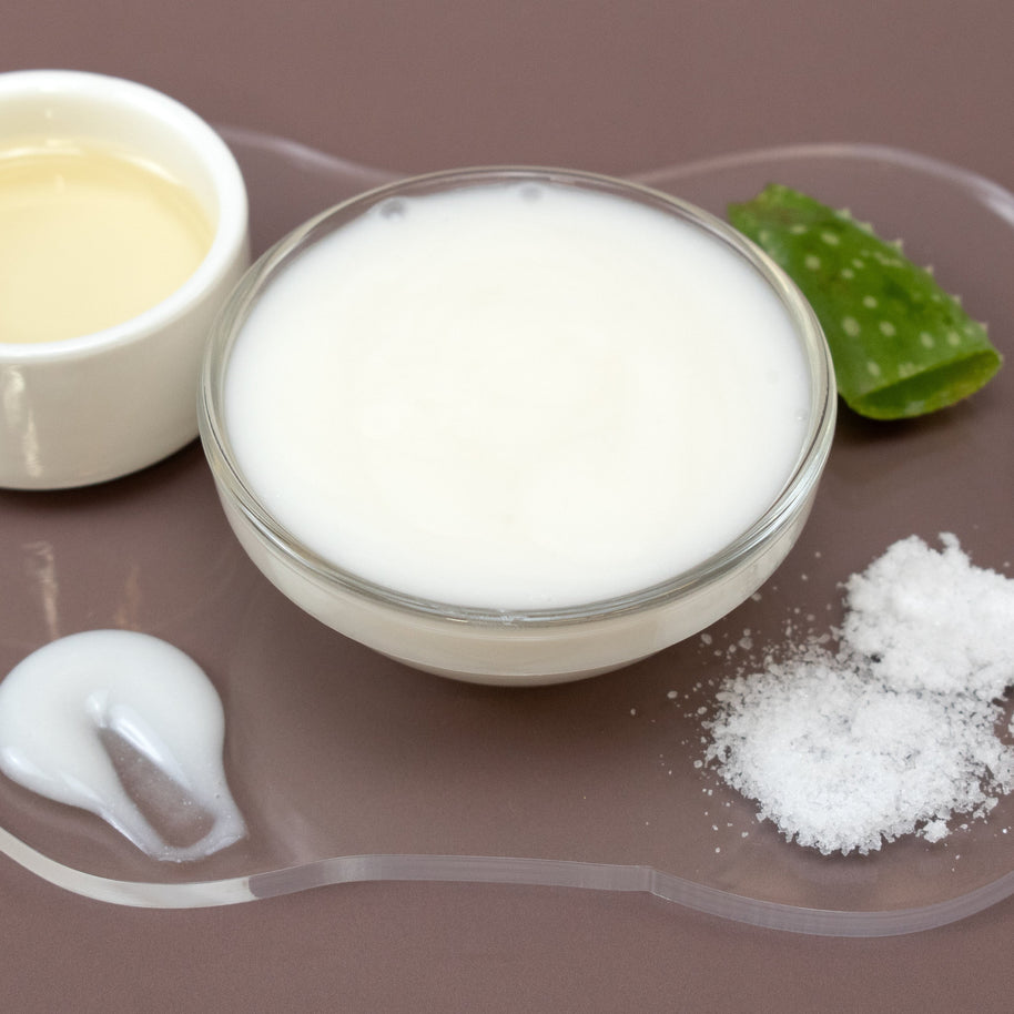 Glass bowl of DMAE & MSM Facial Serum with aloe vera leaf and leaves on a brown background