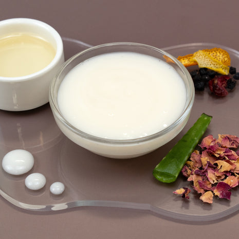Glass bowl of Supreme Facial Serum with a small container of oil, aloe leaf, and dried flowers on a gray surface.