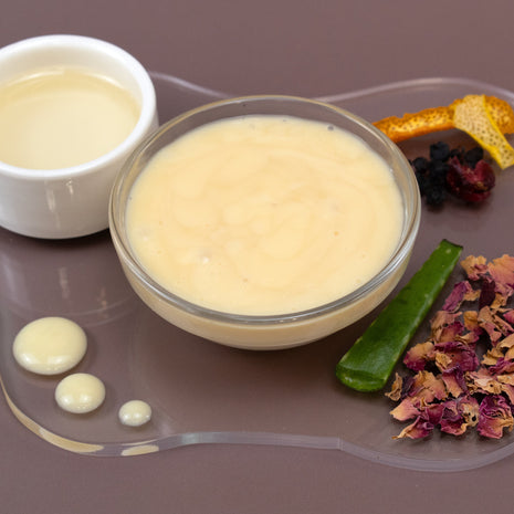 Glass bowl of Ultimate Facial Serum with a white a container of oil, aloe leaf, and dried flowers on a brown surface