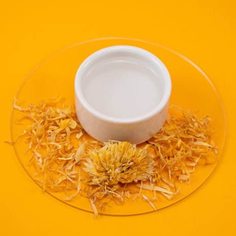 White bowl with Calming Calendula Spray and yellow dried flowers on a clear stand against an orange background