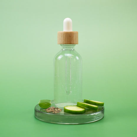 Cucumber Facial Toner in a clear glass bottle with wooden dropper on a green platform with cucumber slices on a solid green background